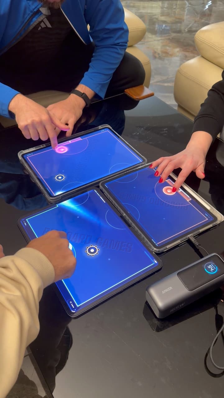 Three tablets pushed together on a coffee table, three players' hands on their mallets; the puck streaks over the seam from one screen to the next.