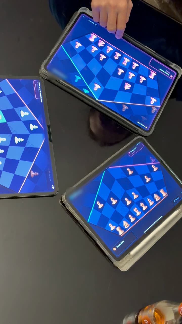 Three tablets on a dark table seen from above, each showing one player's third of a hexagonal three-player chess board, a hand reaching for a piece on the top one.